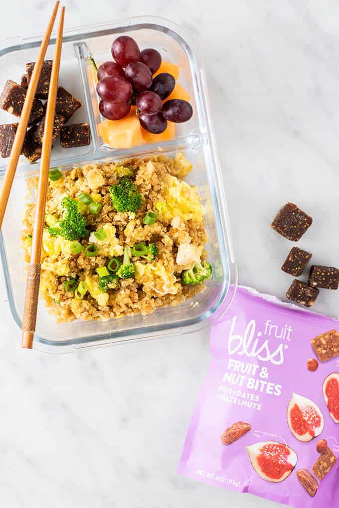 A glass bento box filled with quinoa and broccoli fried rice, some grapes, and a bag of fruit and nut bites.