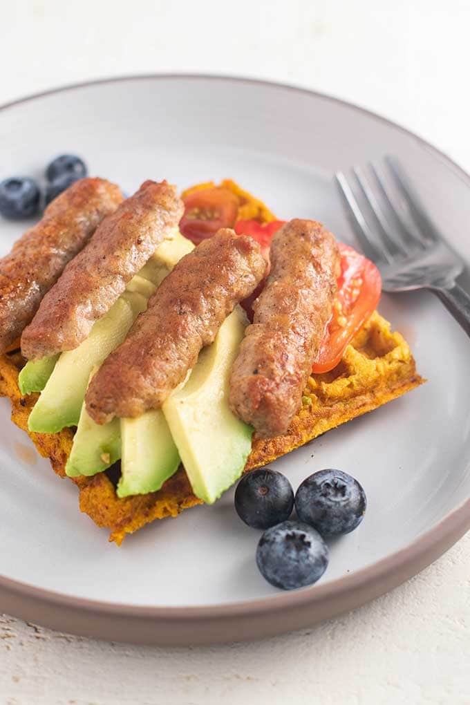 A sweet potato waffle with sliced avocado, sausage, and cherry tomatoes.