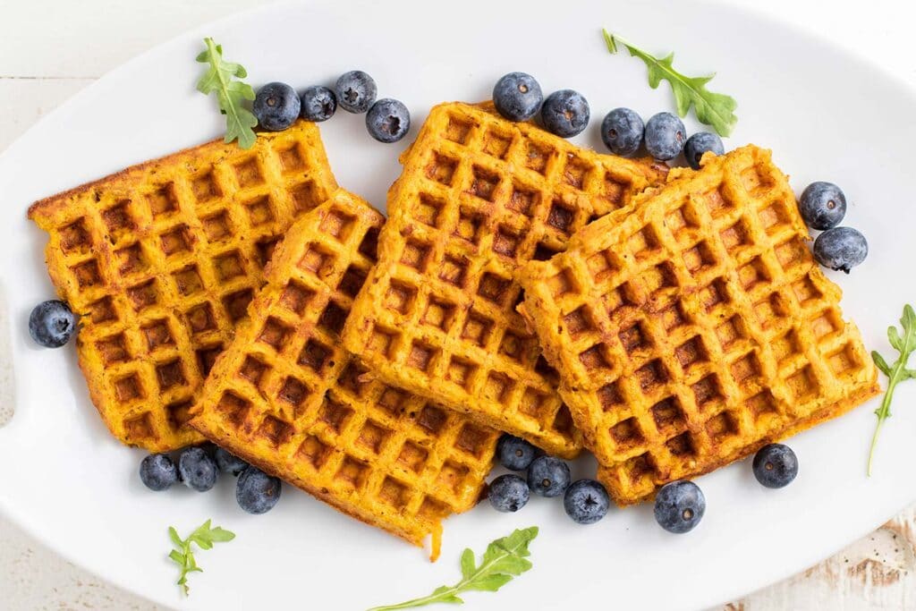 A white plate with sweet potato waffles stacked on it, surrounded by blueberries.
