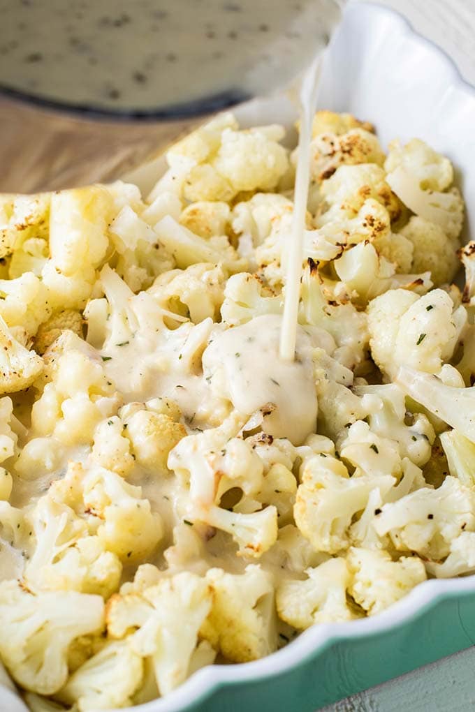 Roasted cauliflower in a baking dish with a cheese sauce being poured over the top.