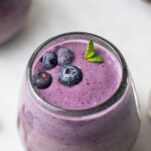 A blueberry smoothie in a clear wine glass garnished with mint and fresh blueberries.