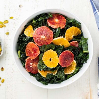 A top down look at a white salad bowl filled with massaged kale, topped with citrus slices, and a bowl of toasted pistachios next to it.