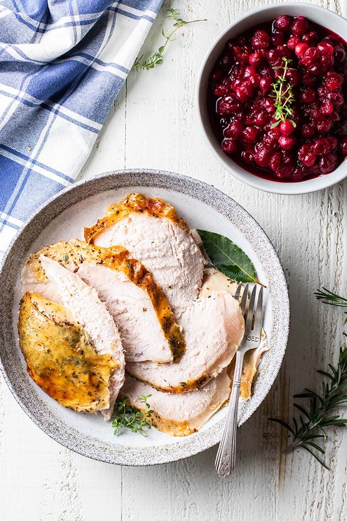 Slices of turkey on a plate garnished with sage leaves next to a bowl of cranberry sauce.