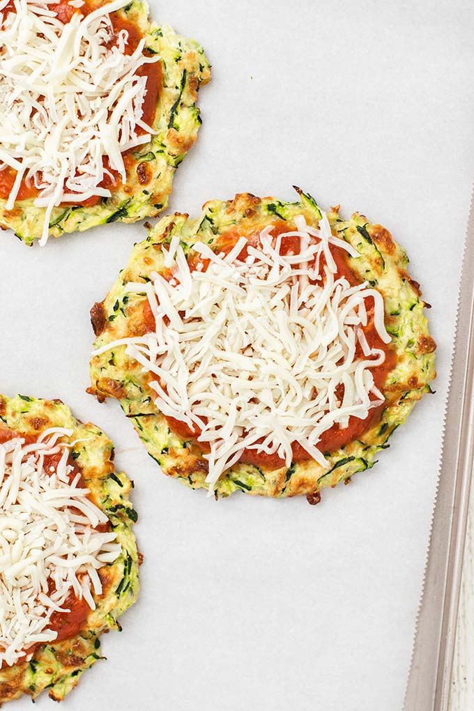 Mini zucchini pizza crusts on a baking tray topped with pizza sauce and mozzarella cheese.