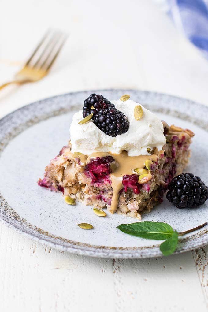 A slice of baked oatmeal on a plate topped with whipped cream and more blackberries.