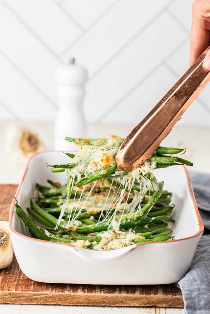 Tongs pulling a serving of garlic green beans from the dish, with strings of cheese pulling from the pan.