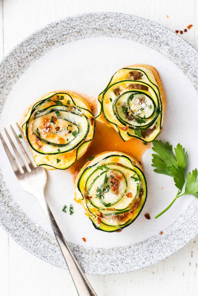 A white plate shown with 3 zucchini roll ups being served with a sprinkle of parmesan cheese on top.