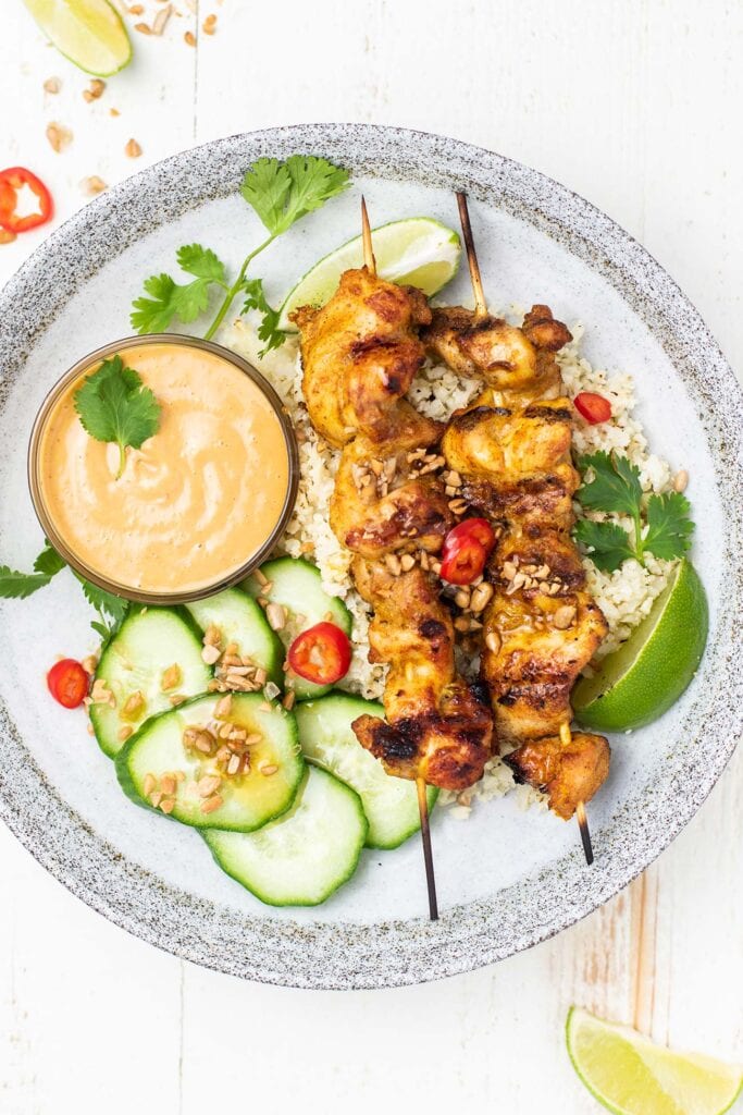 A plate with cauliflower rice, cucumber salad, and 2 chicken satay skewers.