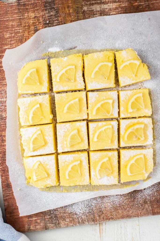 A large cutting board with lemon bars cut into 16 squares and dusted with powdered sugar.
