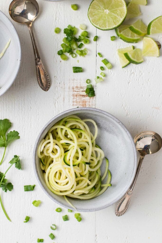 A bowl of zoodles with lime slices, cilantro, and green onions scattered around to show possible garnishes.