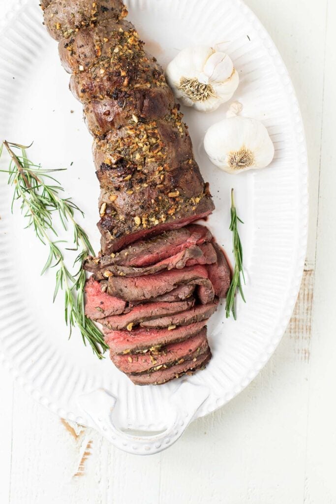 A beef tenderloin roast on a white platter garnished with rosemary.