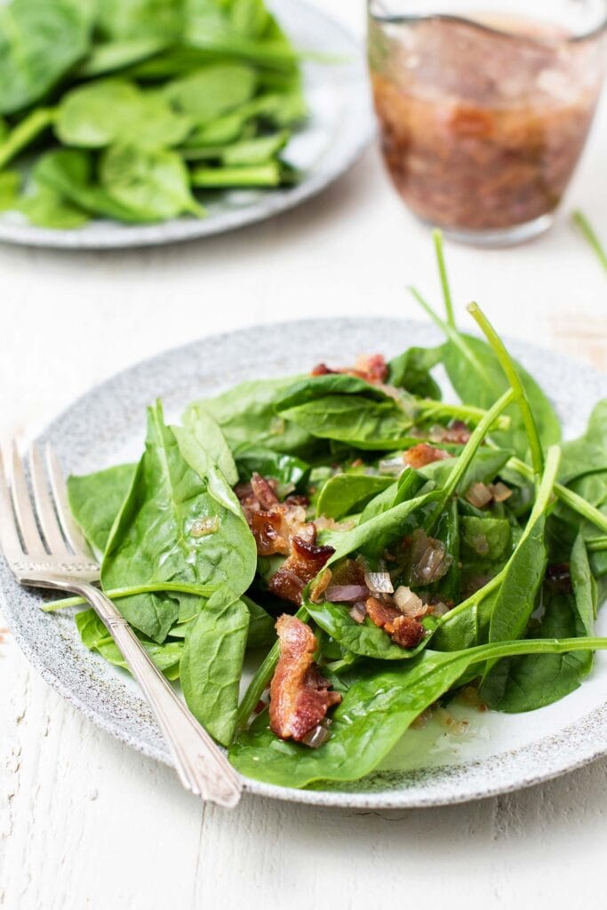 Two plates with spinach salad being topped with hot bacon dressing.
