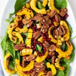 A white platter with a spinach salad topped with delicata squash, pomegranate, and pecans.