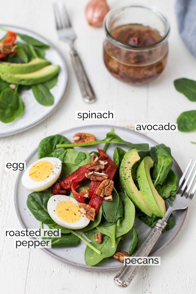 A spinach salad topped with eggs, pecans, roasted red peppers and avocados, shown with labels to describe how to put the salad together.