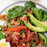 A close up look at a spinach salad topped with eggs, roasted red pepper, avocado, and pecans.