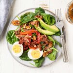 A beautiful salad topped with eggs, avocado, and roasted red peppers, topped with a hot bacon dressing.