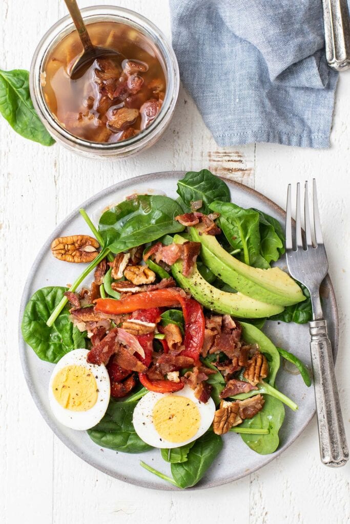 A beautiful salad topped with eggs, avocado, and roasted red peppers, topped with a hot bacon dressing.