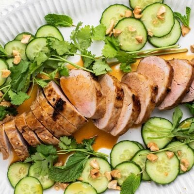 A white platter with a sliced pork tenderloin, garnished with cucumbers, cilantro, mint and cashews.