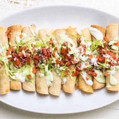 A white plate with a row of taquitos, garnished with salsa and avocado crema.