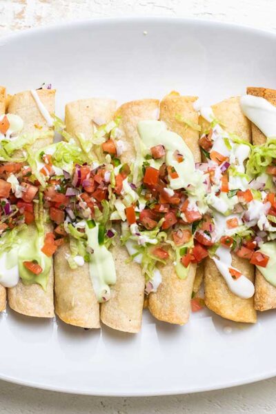 A white plate with a row of taquitos, garnished with salsa and avocado crema.