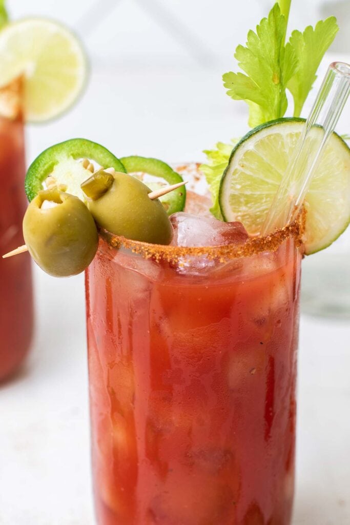 A cocktail shown garnished with olives, lime, and jalapeno, and a salted rim.