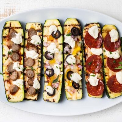 6 grilled zucchini pizza boats lined up on a white serving platter.