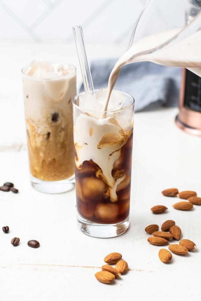 A pitcher pouring almond milk creamer into a glass of iced coffee.