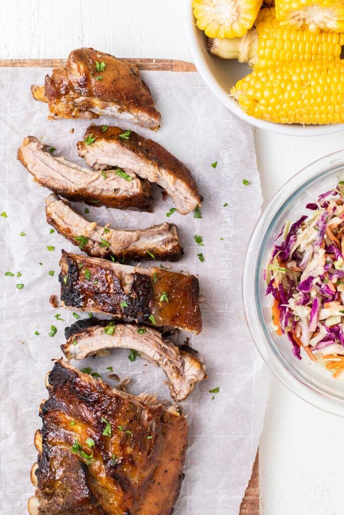 A rack of ribs shown being cut next to corn on the cob and cole slaw.
