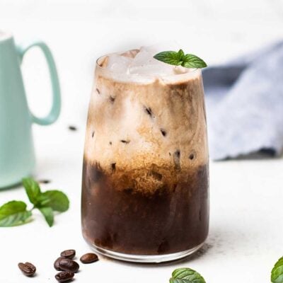 A coconut milk iced mocha with peppermint shown in a glass with mint leaves and coffee beans.