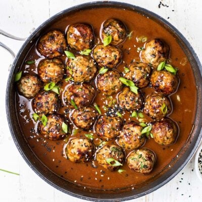 Asian Pork Meatballs shown in a spicy sauce garnished with sesame seeds and green onions.