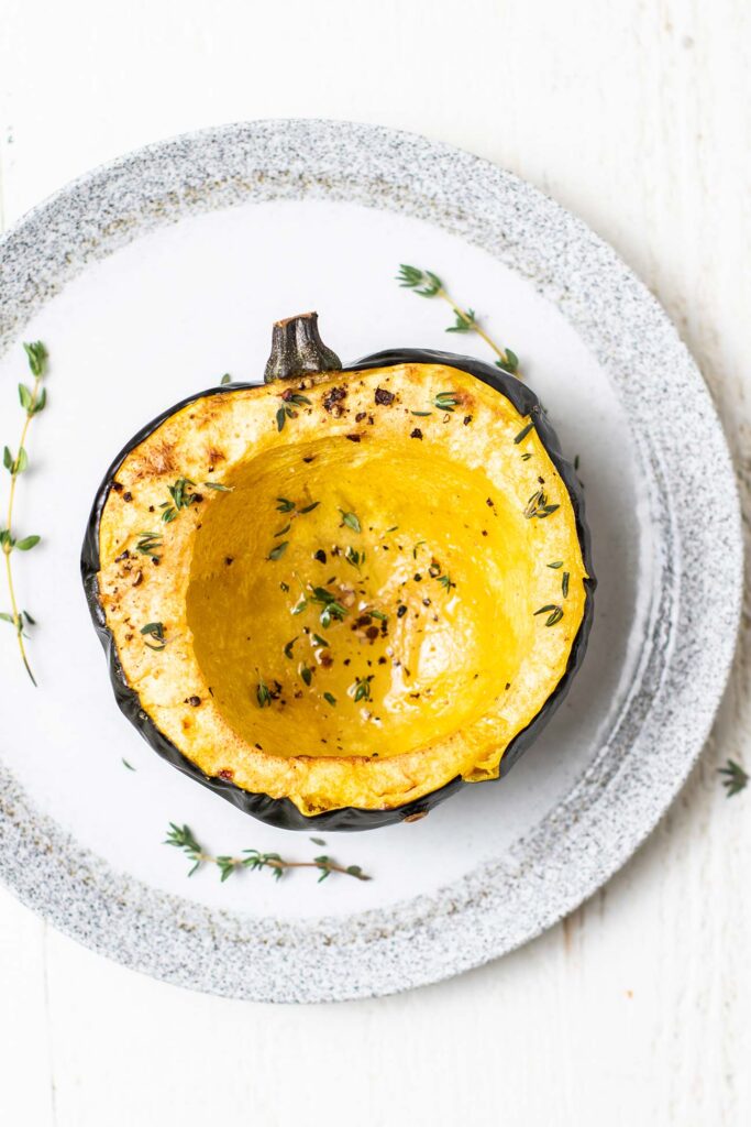 Half of a roasted acorn squash shown on a plate garnished with fresh thyme.