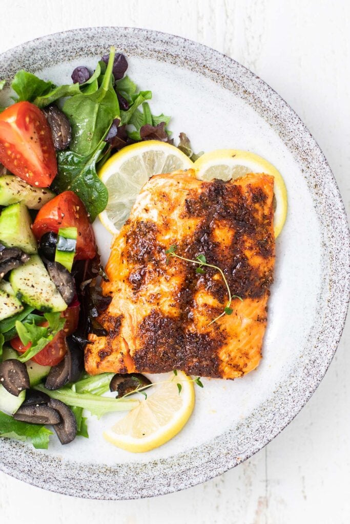 A piece of blackened salmon shown on a plate with salad and lemon slices.