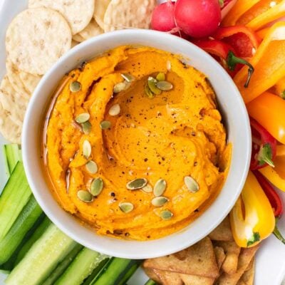 A close up look at a dish of carrot hummus surrounded by crackers and bright veggie sticks.