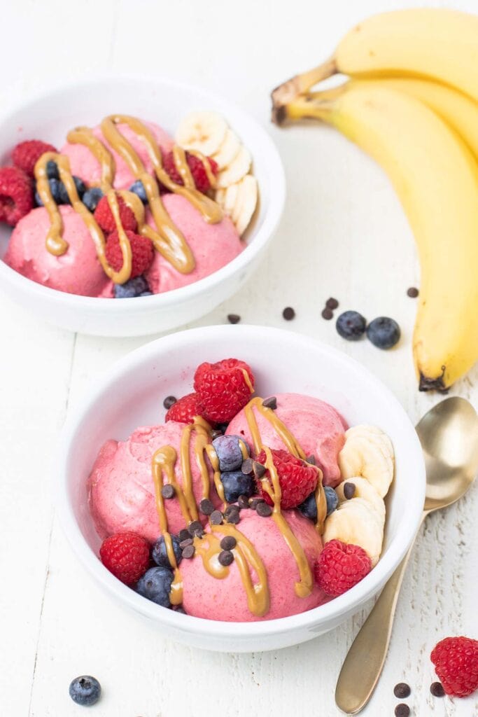 Two white bowls filled with 3 scoops of bright pink breakfast ice cream, topped with berries and nut butter.