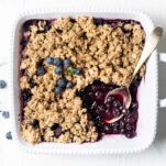 A blueberry crisp in a white baking dish, shown with a gold spoon scooping some out.