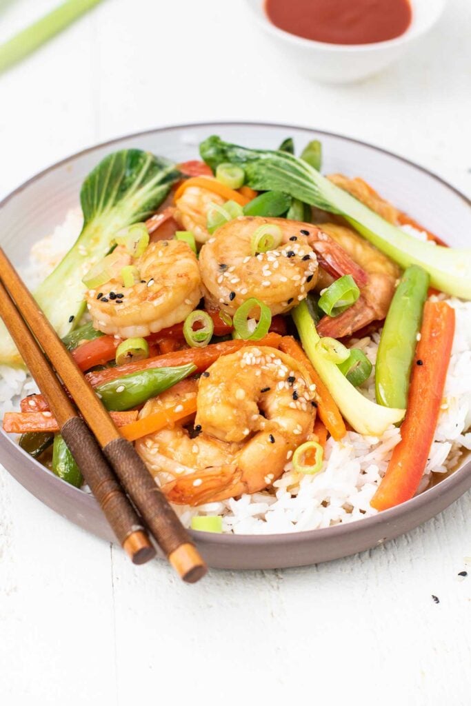 A white plate with a serving of rice topped with a shrimp stir fry, with chopsticks on the side.