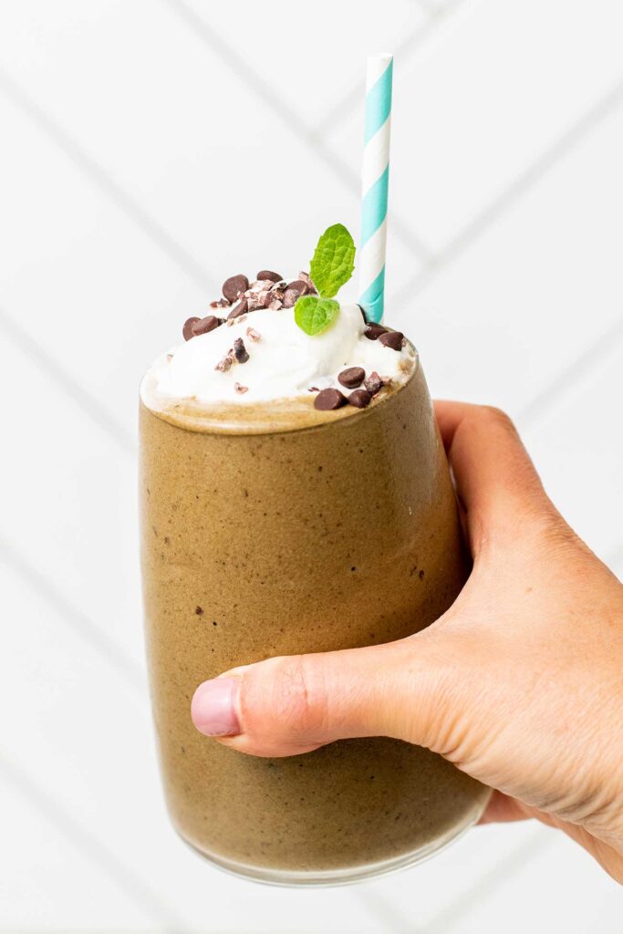 A hand holding a glass filled with a mocha green smoothie topped with whipped cream and cacao nibs.