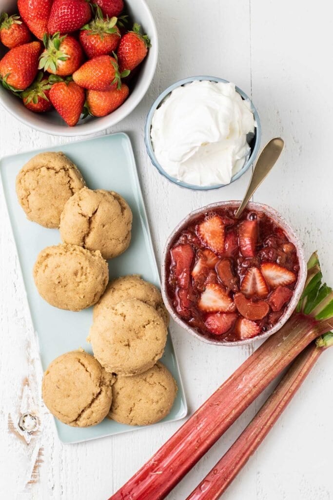 Shortcakes shown on a platter next to a bowl of strawberries, strawberry rhubarb compote, and whipped cream.