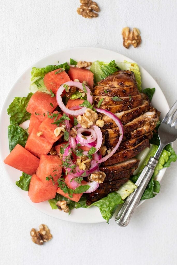 A white plate with lettuce, watermelon, and balsamic chicken.