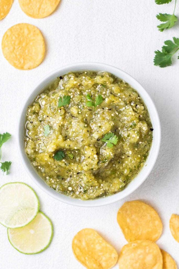 A bowl of roasted tomatillo salsa with tortilla chips and lime surrounding it.