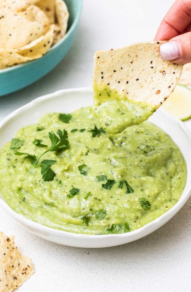 A bowl of creamy avocado salsa verde shown with a chip dipping in to it.