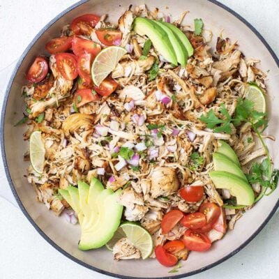 A white cast iron pan filled with tender chunks of shredded chicken, topped with cilantro, onions, tomatoes and avocado.