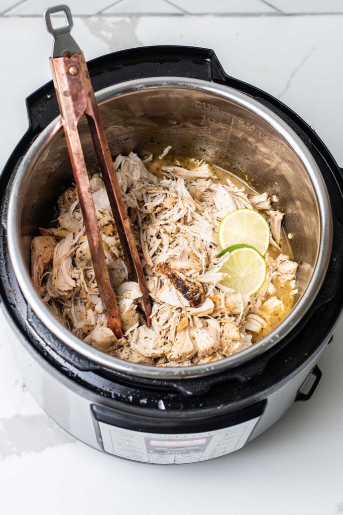 Mexican pulled chicken in an Instant Pot shown with tongs pulling some out.