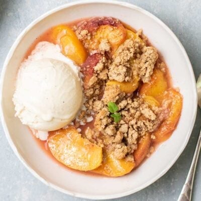 A white bowl with peach crumble and a big scoop of vanilla ice cream.