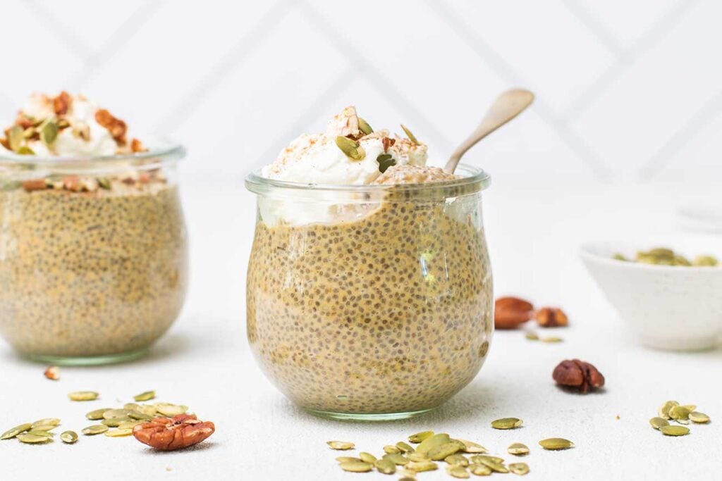 Two jars of pumpkin spice chia seed pudding topped with whipped cream, shown with pecans and pumpkin seeds scattered on the countertop.