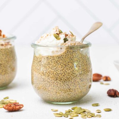 Two jars of pumpkin spice chia seed pudding topped with whipped cream, shown with pecans and pumpkin seeds scattered on the countertop.