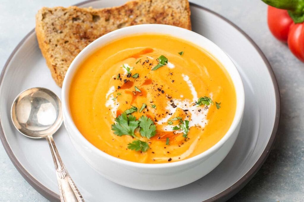 A bowl of sweet potato roasted red pepper soup shown with a slice of buckwheat bread.