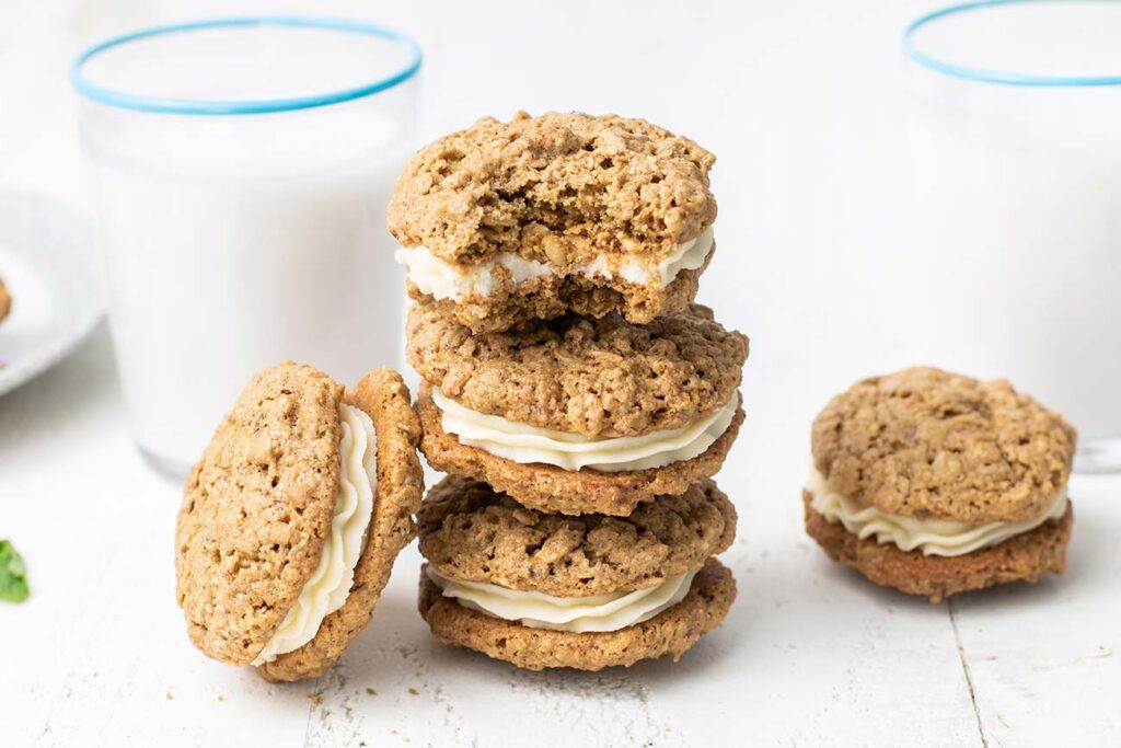 A stack of oatmeal cream pies shown in front of glasses of almond milk.