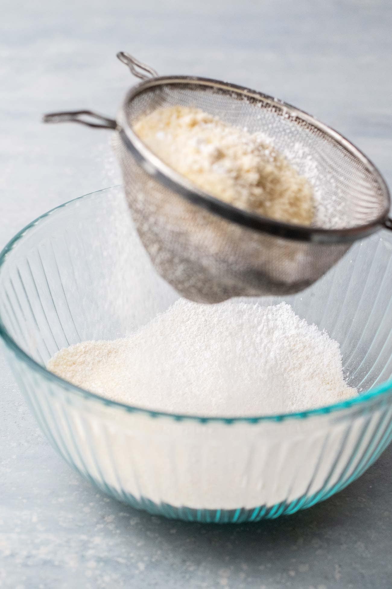 A sifter with almond flour and powdered sugar, sifting into a glass bowl.