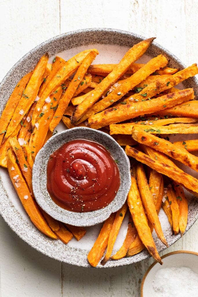A plate with sweet potato fries shown surrounding a dish of ketchup.